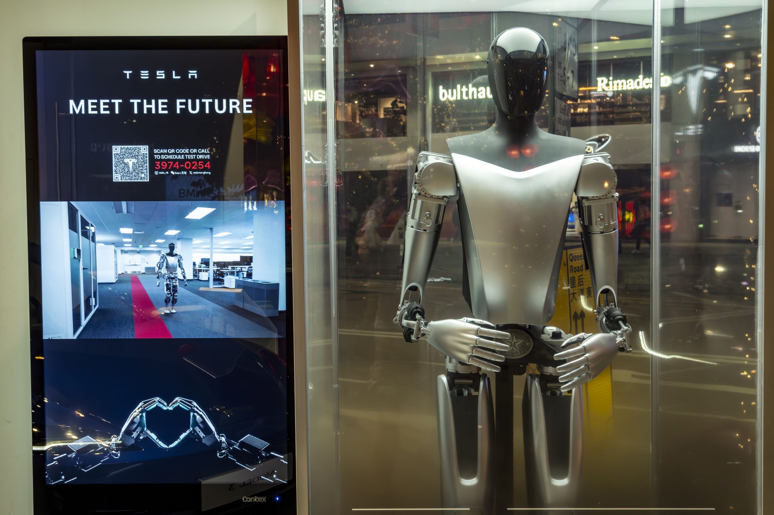 The Optimus robot, which looks like a metal humanoid with head and arms, stands next to a screen at a Tesla event.