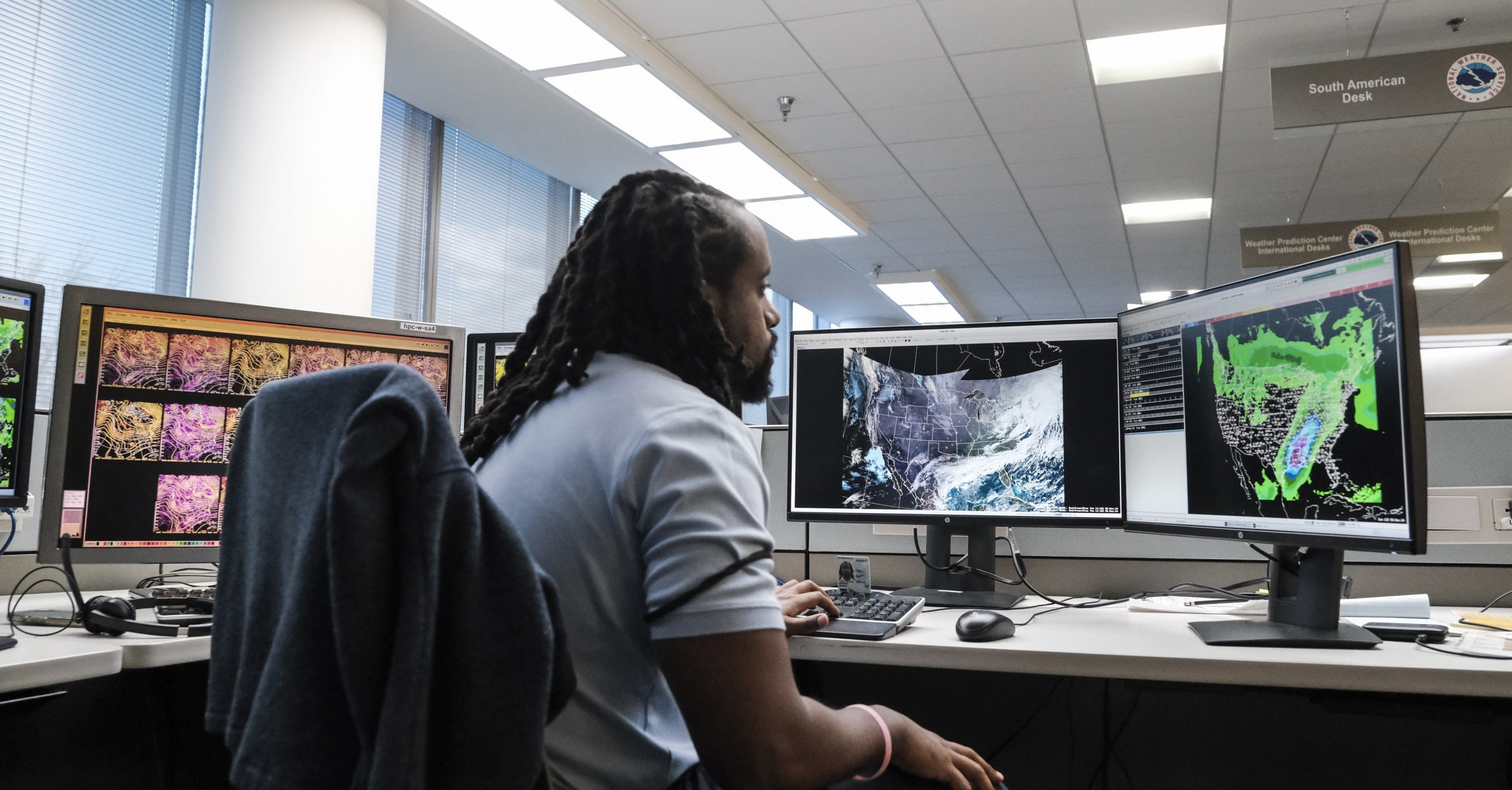 A man at a desk looks at several computer screens with maps of the US and weather blobs over the map. A man at a desk looks at several computer screens with maps of the US and weather blobs over the map.