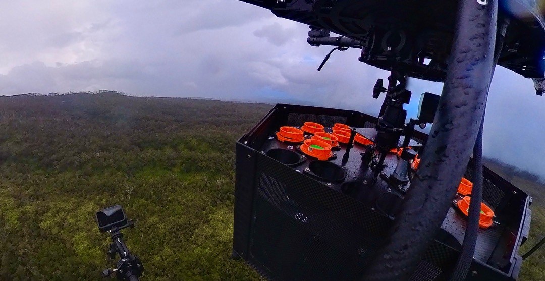 A drone’s eye view of its own frame, with a black box containing orange capsules attached to it, hovering over a forest that stretches out green to the horizon. A drone’s eye view of its own frame, with a black box containing orange capsules attached to it, hovering over a forest that stretches out green to the horizon.
