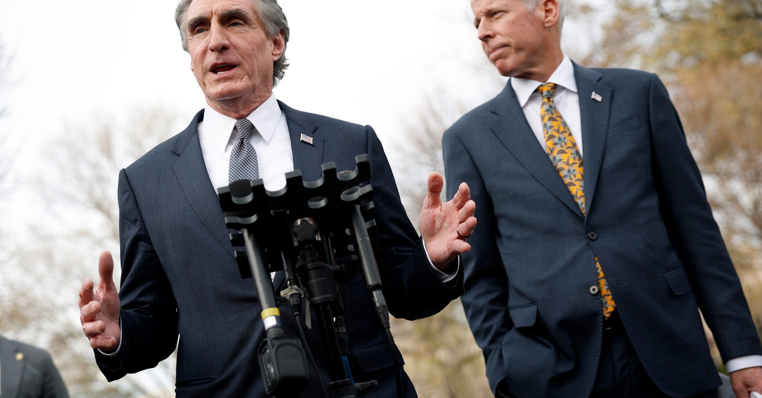 Seen from a low angle below, Doug Burgum, in a navy suit, talks into a gaggle of press microphones, with another man in a navy suit standing next to him.