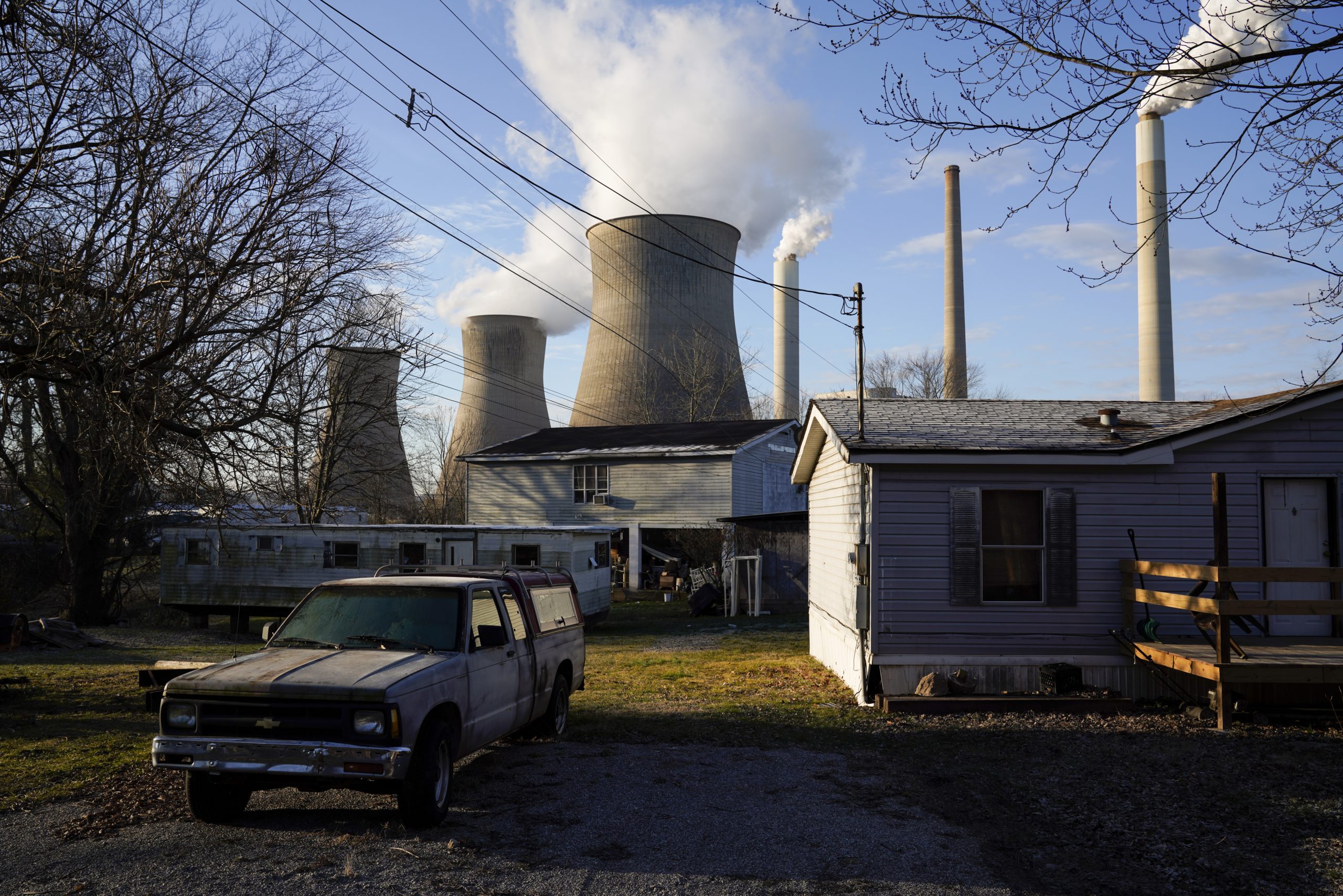 A coal-fired power plant emits plumes of smoke from multiple towers; in the foreground are houses and a truck.