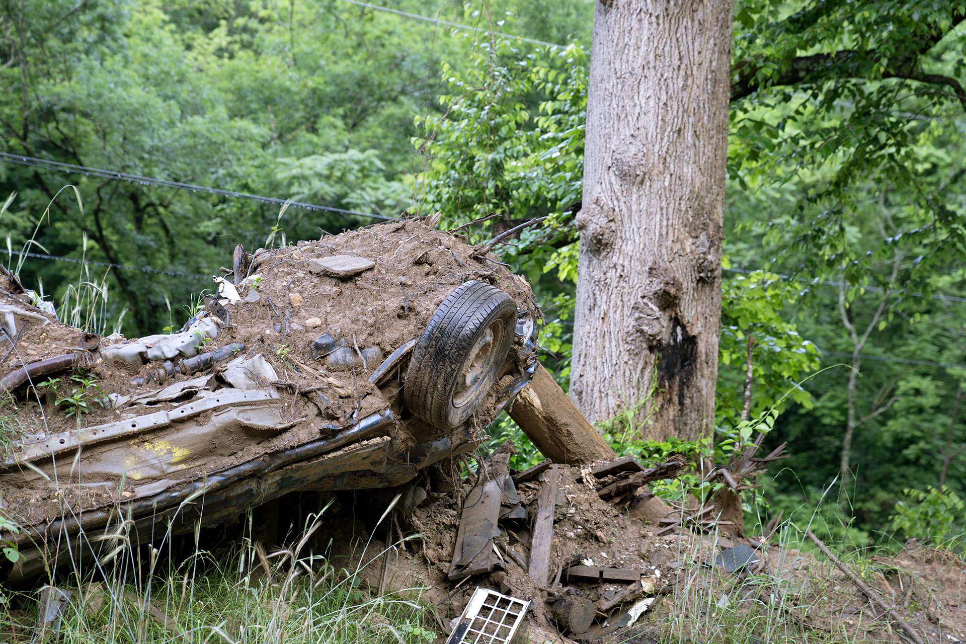 A car is severely damaged, covered in mud and grass, and turned upside down in a forest.