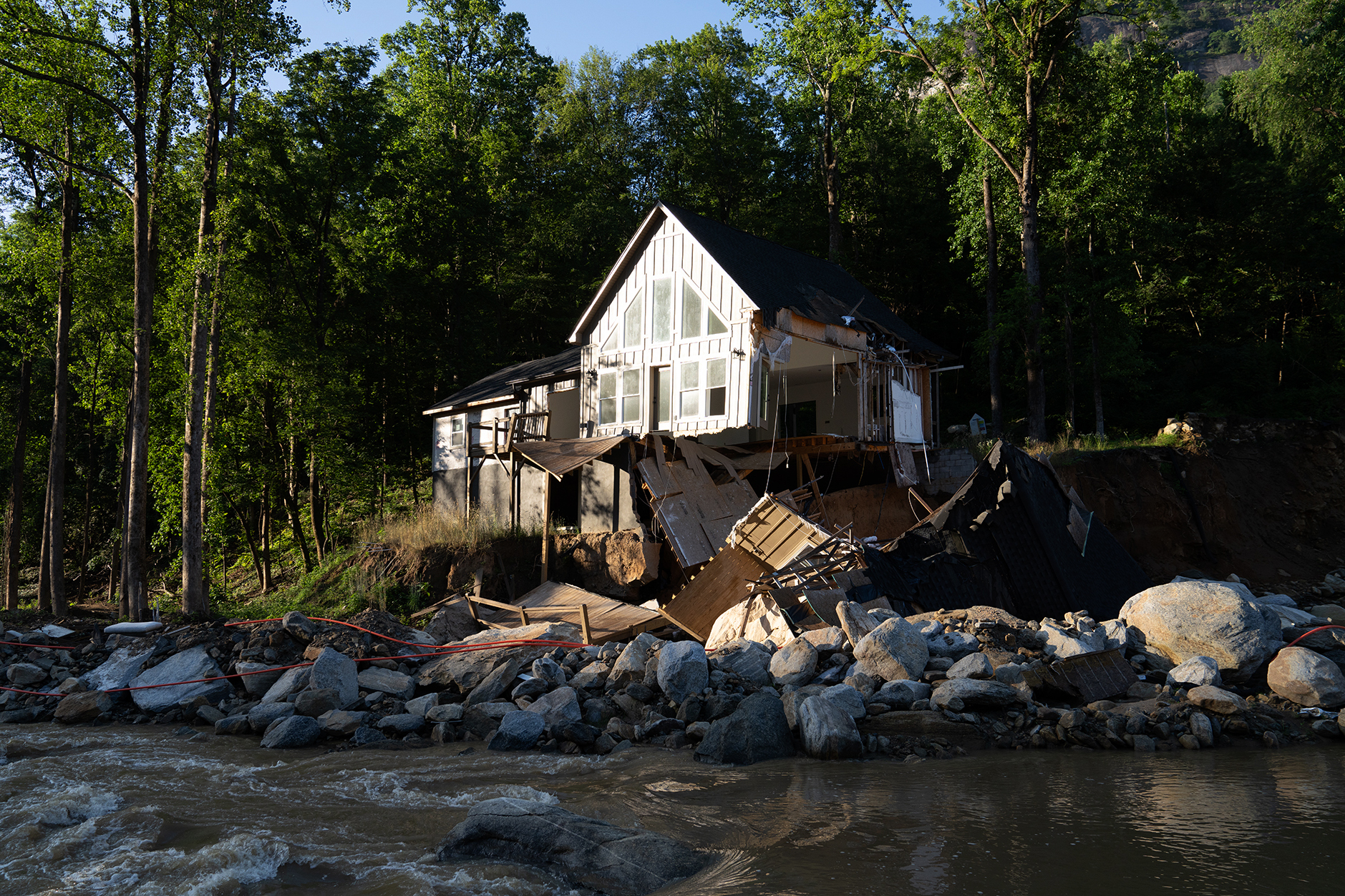 A home backed up to a forest with severe hurricane damage. Large pieces of the home have been ripped away and are scattered on rocks and water below.