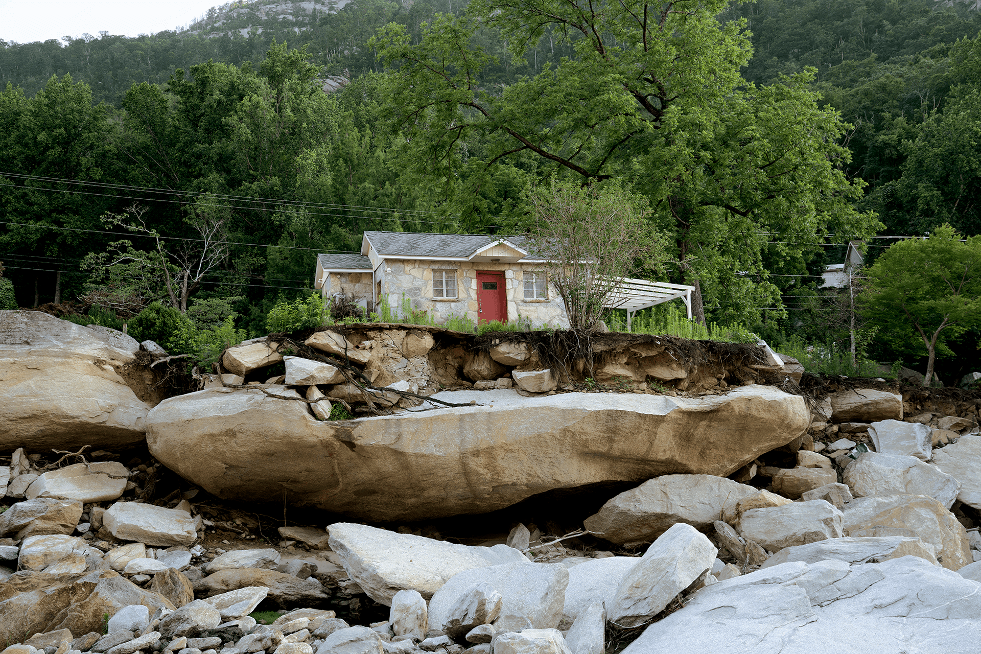 A home abutting a lush forested area sits behind a dry riverbed with large rocks.