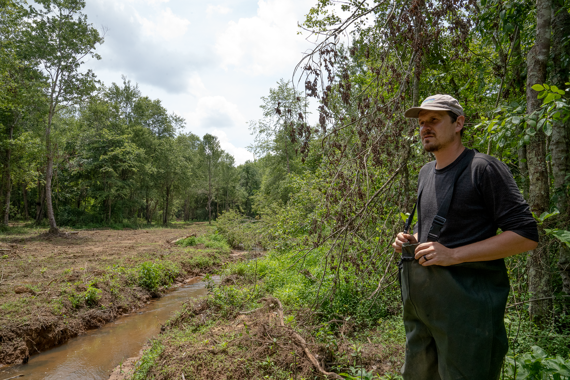 A man wearing waders stands at the edge of a river, surrounded by trees