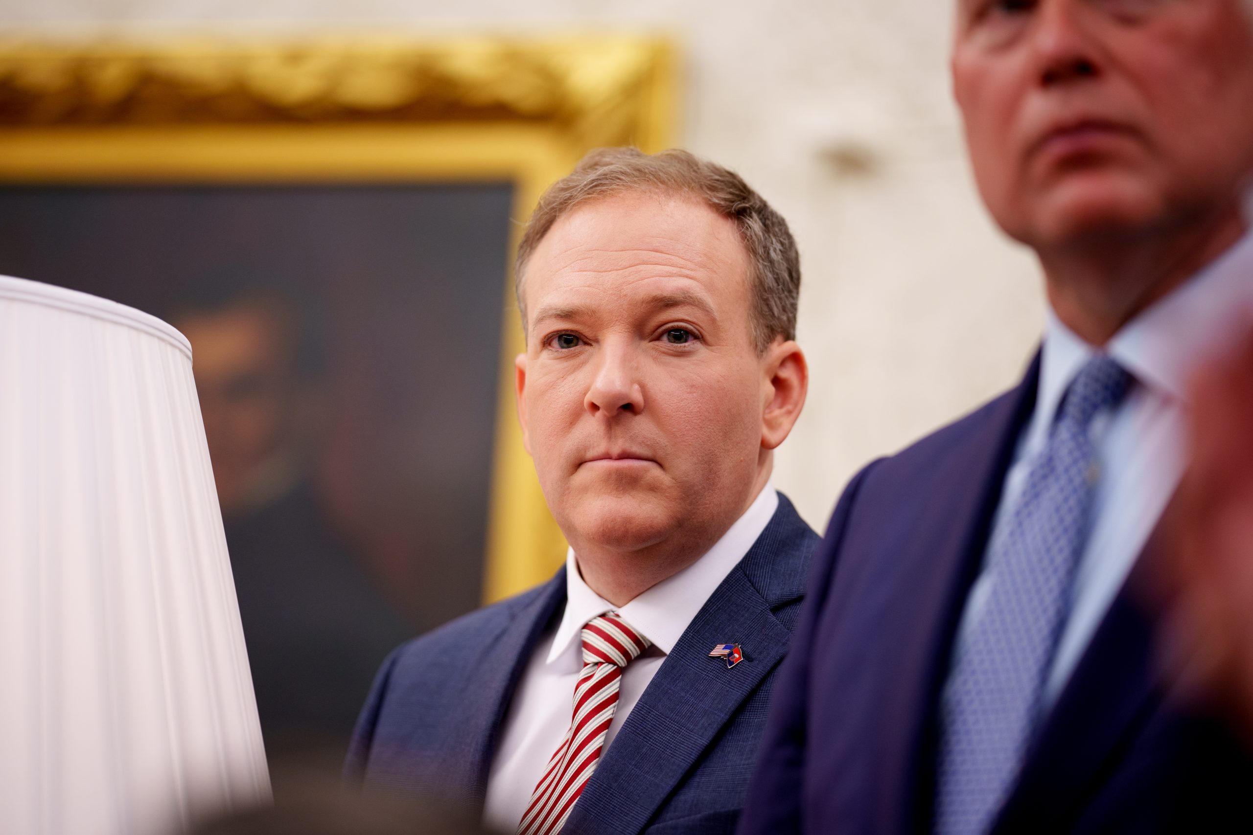 Lee Zeldin, wearing a navy suit and striped tie, is seen in the Oval Office; another man wearing a suit is partially visible to his right.