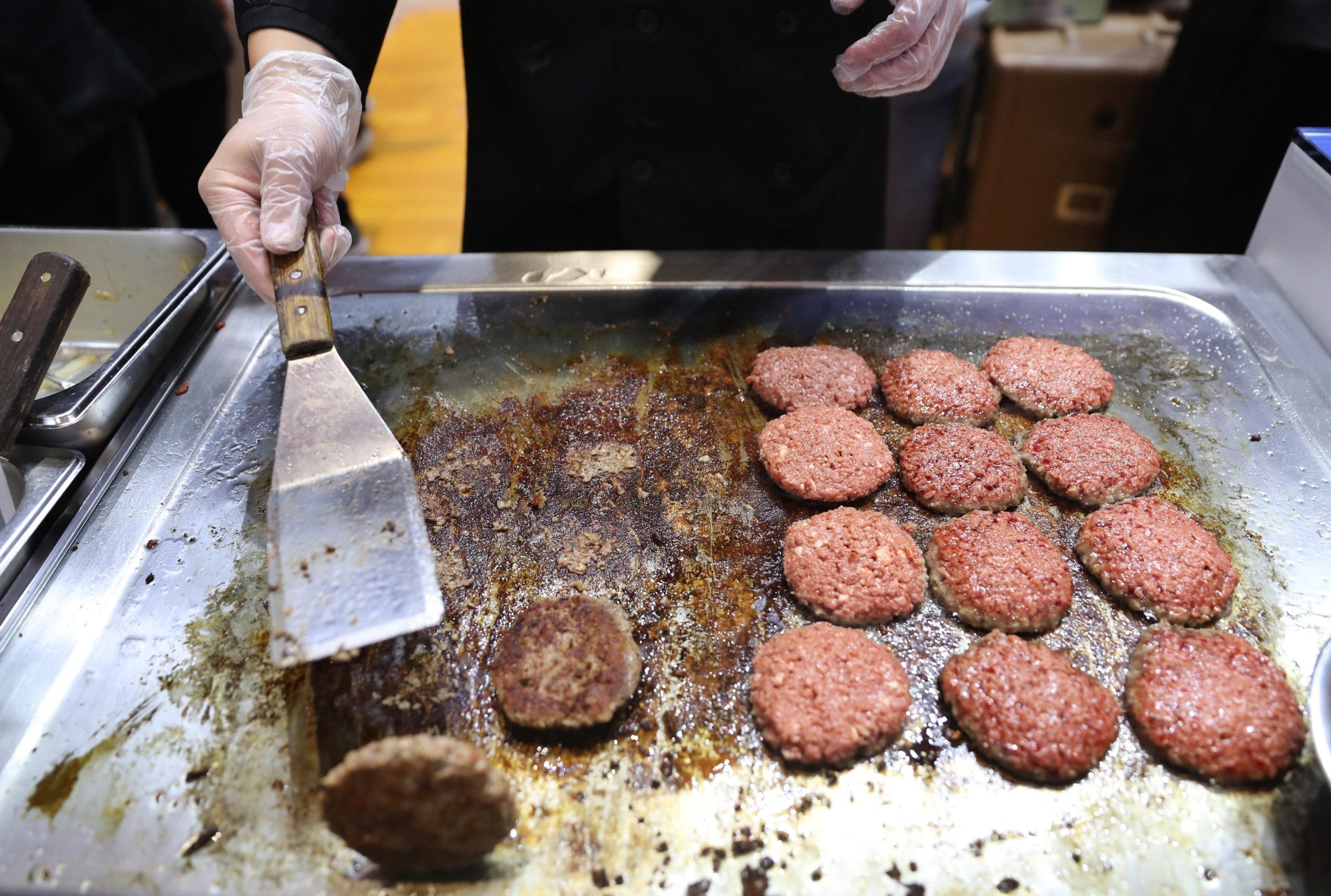 Around a dozen Impossible burgers cooking on a grill. A person is flipping one. 