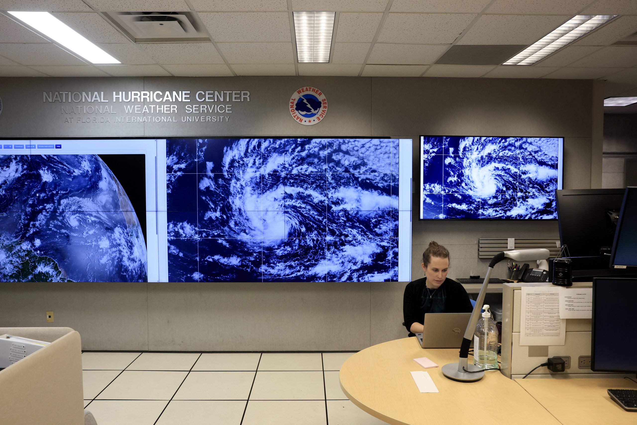 A NOAA hurricane specialist tracks Tropical Storm Erin as it strengthens into the season’s first major hurricane. She sits at a computer in front of three large screens showing the latest satellite images of the storm.