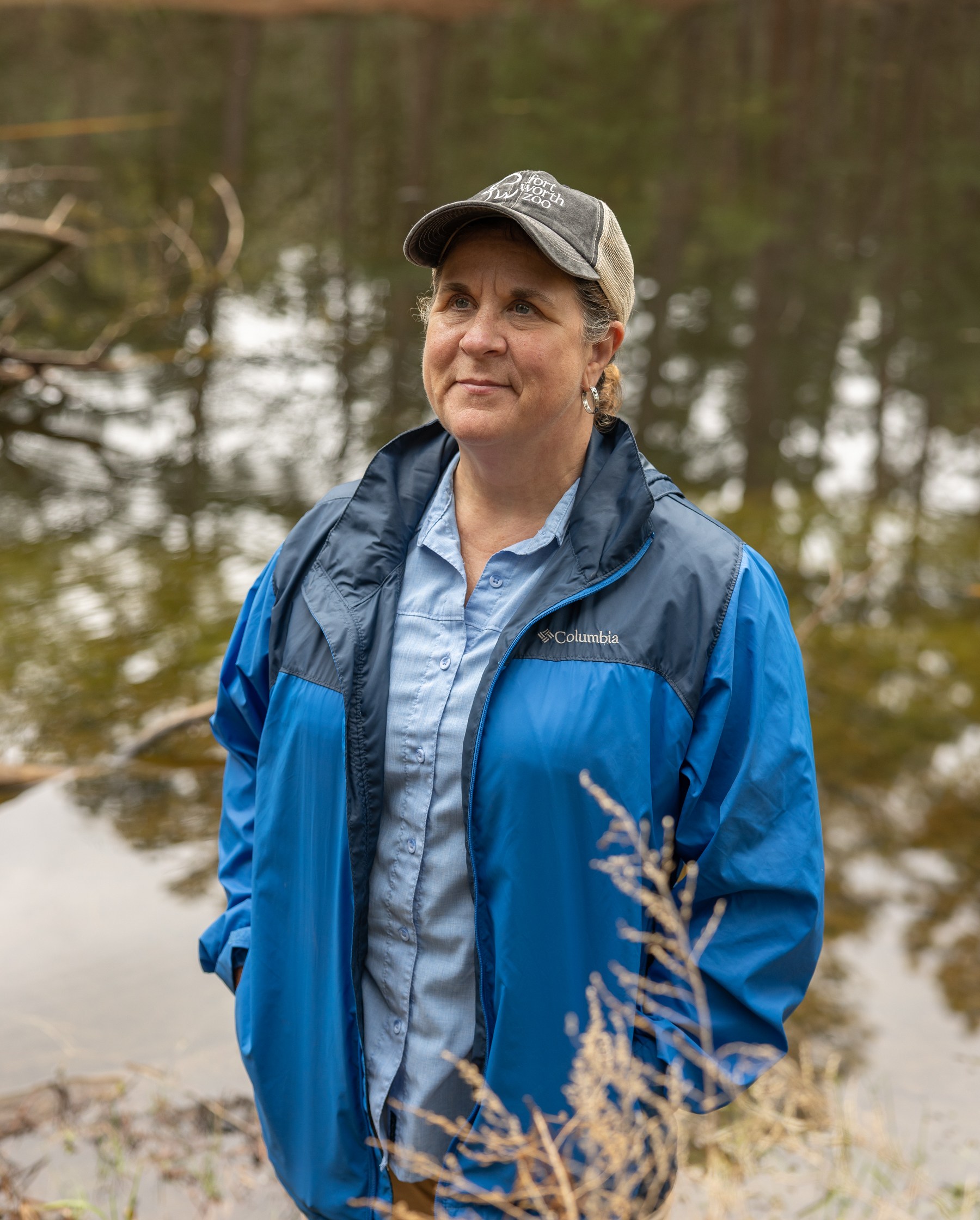 A middle-aged woman wearing a blue jacket and hat stands near a pond A middle-aged woman wearing a blue jacket and hat stands near a pond